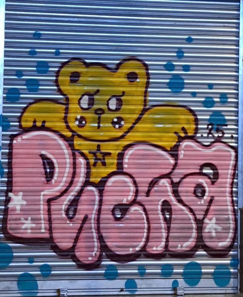 Graffiti de un oso de peluche amarillo con detalles distintivos, como un ojo en 'X' y una estrella en el pecho, pintado sobre la persiana metálica de la tienda "Shopping Barcelona" en Carrer Hospital, Barcelona. El fondo de la persiana tiene lunares azules